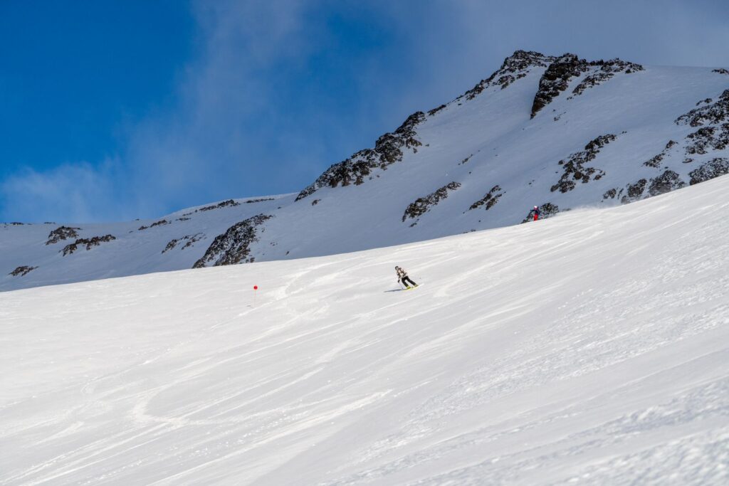 Skier descending a broad snowy slope beneath a rocky mountain ridge and blue sky